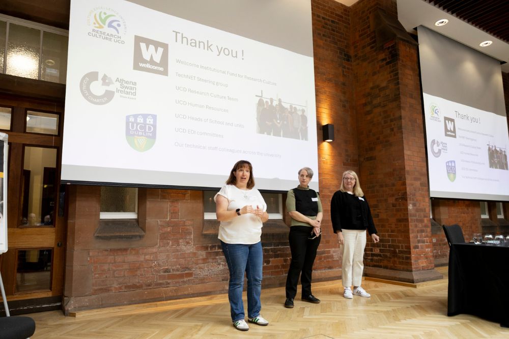 Three female speakers standing at the front of a large red brick hall with parque flooring speaking in front of a large overhead screen displaying the words 'Thank you' and the logos of Research culture UCD, Wellcome, Athena Swan Ireland and UCD